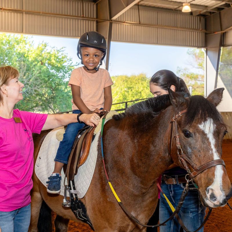 Young girl smiling and sitting on top of a horse in the Equest arena while a staff member supervises