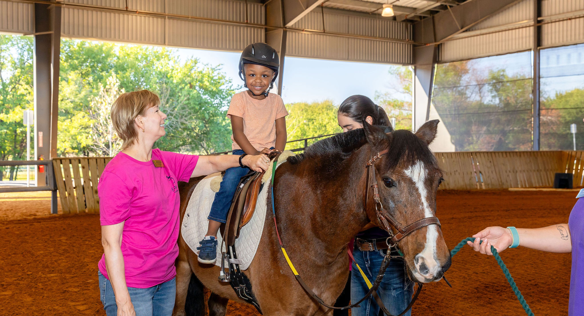 Young girl smiling and sitting on top of a horse in the Equest arena while a staff member supervises